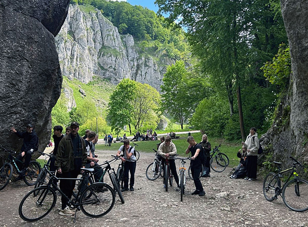 Grupa uczniów z rowerami stoi na kamienistej drodze między wysokimi, szarymi skałami w zielonej dolinie Ojcowskiego Parku Narodowego. W tle widać strome, jasne skały, drzewa z bujną zielenią i błękitne niebo z białymi chmurami. Na pierwszym planie, stoi kilku uczniów z rowerami, niektórzy patrzą w stronę aparatu, a inni zajmują się swoimi rowerami, opierając je o siebie lub trzymając za kierownice. Po prawej stronie zdjęcia siedzi lub stoi kilka osób, które odpoczywają, a obok nich oparte są rowery. W głębi zdjęcia, za grupą, widać jeszcze więcej osób siedzących na trawie lub przechodzących.