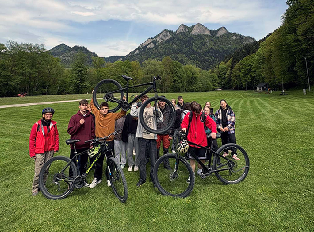 Grupa uczniów stoi z rowerami na zielonej łące w Pieninach, na tle gór Trzy Korony. Jedna osoba trzyma rower uniesiony w górze. W tle widać lasy i charakterystyczne szczyty górskie, a nad nimi błękitne niebo z białymi chmurami. Uczniowie są uśmiechnięci, ubrani w sportowe stroje, gotowi na dalszą rowerową trasę w otoczeniu przyrody.