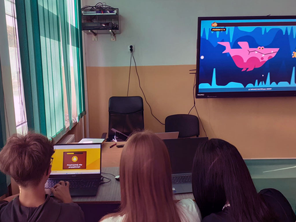 Na zdjęciu widać trójkę uczniów siedzących tyłem przy biurku w klasie. Pracują na laptopach, które mają przed sobą. Jeden z laptopów pokazuje planszę z napisem „Przygotuj się do gry”, a na drugim widać ekran startowy. Przed uczniami, na ścianie, zawieszony jest duży monitor, na którym wyświetlona jest kolorowa gra edukacyjna z różowym rekinem i niebieskim morskim tłem. Z lewej strony zdjęcia widać okno z zielonymi żaluzjami, przez które wpada światło dzienne. Sala jest jasna, z żółto-białymi ścianami. Uczniowie uczestniczą w zajęciach z wykorzystaniem nowych technologii, ucząc się poprzez gry edukacyjne i interaktywne materiały multimedialne.