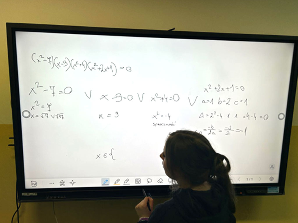 Na zdjęciu widać uczennicę stojącą tyłem przy dużym monitorze interaktywnym w sali lekcyjnej. Ekran jest biały, a na nim widoczne są czarne odręczne zapisy równań i działań matematycznych dotyczących rozwiązywania równań kwadratowych. Dziewczynka ma spięte włosy i ciemną bluzę, patrzy na ekran i trzyma w ręce pisak, którym może pisać lub zaznaczać treści na tablicy interaktywnej. Zdjęcie pokazuje, jak uczniowie wykorzystują nowoczesne technologie na lekcji matematyki,  rozwiązując zadania przy pomocy tablicy multimedialnej.