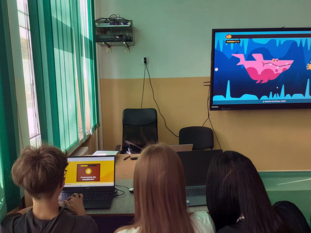 Na zdjęciu widać troje uczniów siedzących przy jednym stole w sali lekcyjnej. Uczniowie siedzą tyłem, widać ich głowy i ramiona. Przed uczniami leżą dwa laptopy. Na laptopie po lewej stronie widać żółty ekran z napisem „Przygotowuję się, przygotowuję…”, co wskazuje, że trwa ładowanie gry lub programu. Na ścianie przed uczniami wisi duży monitor interaktywny, na którym wyświetlona jest kolorowa grafika z różowym rekinem na niebieskim tle, co sugeruje, że trwa gra edukacyjna lub interaktywne ćwiczenie. Po lewej stronie zdjęcia znajduje się okno z zielonymi pionowymi żaluzjami, przez które wpada światło dzienne. W tle widać biurko nauczyciela z krzesłem oraz półkę z urządzeniami elektronicznymi.