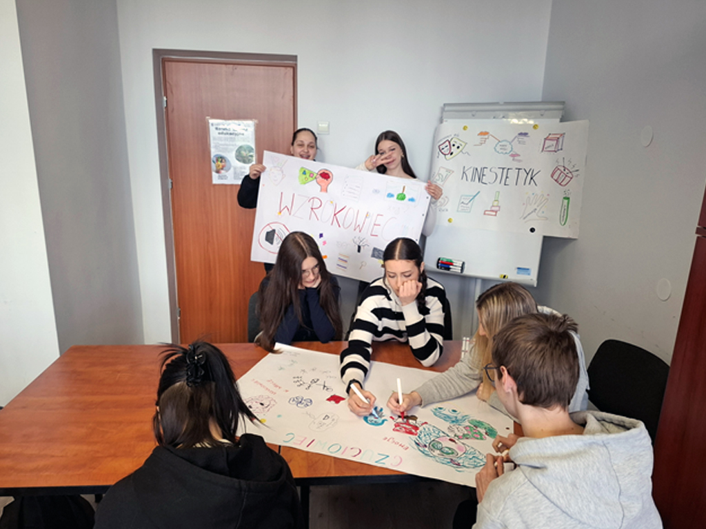 Na zdjęciu widać grupę uczniów podczas zajęć warsztatowych w sali lekcyjnej. Po lewej stronie dwie uczennice stoją i prezentują plakat z kolorowymi rysunkami i napisem „wzrokowiec”. Po ich lewej stronie stoi tablica suchościeralna, a na niej umieszczony jest kolorowy plakat. Na środku plakatu umieszczony jest napis „kinestetyk”, obok napisu są różne kolorowe obrazki.  Przy stoliku siedzi pięciu uczniów. Maja przed sobą duży arkusz papieru, w dłoniach trzymają flamastry, którymi rysują i piszą. Na górze nowo tworzonego plakatu widnieje napis „czuciowiec”. W tle widać drzwi a na nich naklejony plakat projektu Równe szanse edukacyjne.