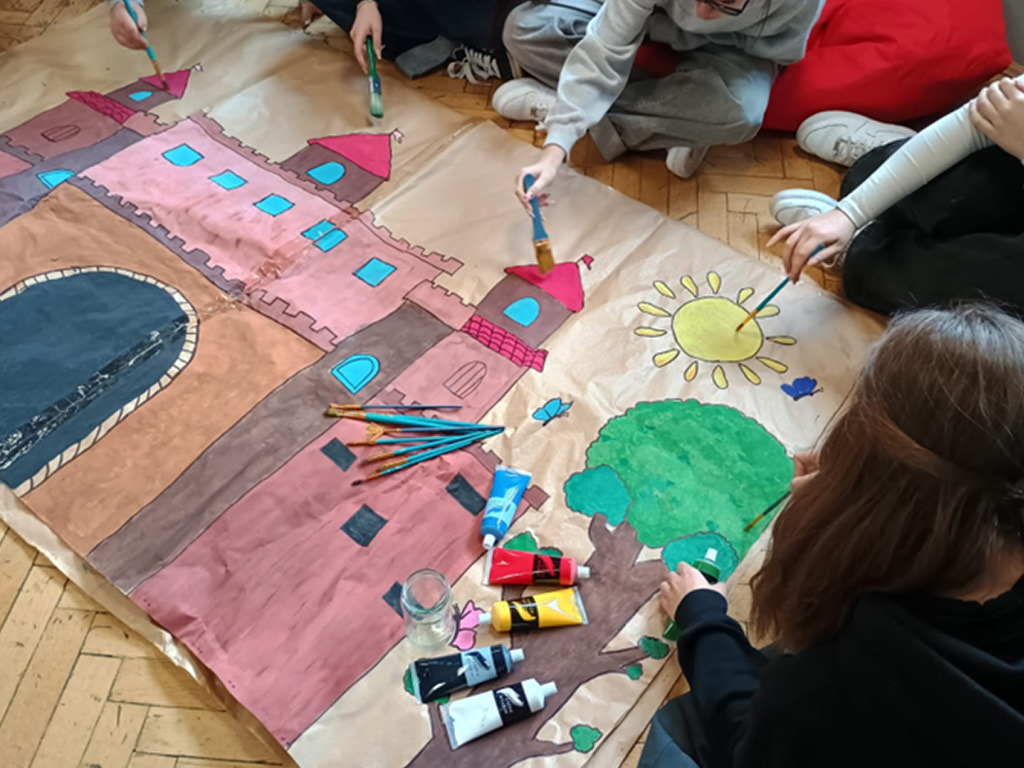 Na zdjęciu widać grupę uczniów, które siedzą na drewnianej podłodze i malują dużą pracę plastyczną na papierze pakowym.  Praca przedstawia różowy zamek z wieżami o czerwonych dachach i błękitnych oknach, dużą czarną bramę, zielone drzewo z brązowym pniem, żółte słońce z promieniami oraz dwa niebieskie motyle.  Uczniowie używają pędzli i kolorowych farb, które leżą w tubkach na środku pracy. Pracują razem, każdy maluje inny fragment obrazka.