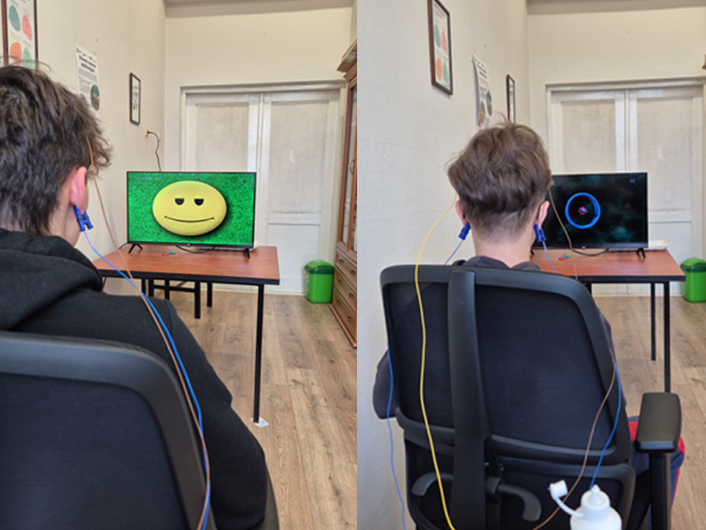 Na zdjęciu z lewej strony, widać ucznia siedzącego na krześle tyłem do obiektywu. Na głowie ma założone elektrody podłączone do urządzenia do biofeedbacku. Przed nim stoi stół, na którym znajduje się monitor, na którym na zielonym tle wyświetla się żółta buźka emotikona z czarnymi oczami i neutralną miną z lekko opuszczonymi kącikami ust, co sprawia, że wygląda na niezbyt uśmiechniętą. Pokój jest jasny, przed uczniem widać białe drzwi, a przed nimi zielony kosz na śmieci. Po lewej stronie na ścianie wiszą obrazki i plakat „Równe szanse edukacyjne”,  a po prawej stoi szafa. Zdjęcie z prawej strony przedstawia ten sam pokój. Widać innego ucznia, który siedzi na krześle  tyłem do obiektywu. Na głowie ma założone elektrody podłączone do urządzenia do biofeedbacku. Przed nim stoi stół, na którym znajduje się monitor. Na czarnym tle monitora wyświetla się niebieski wykres kołowy.  