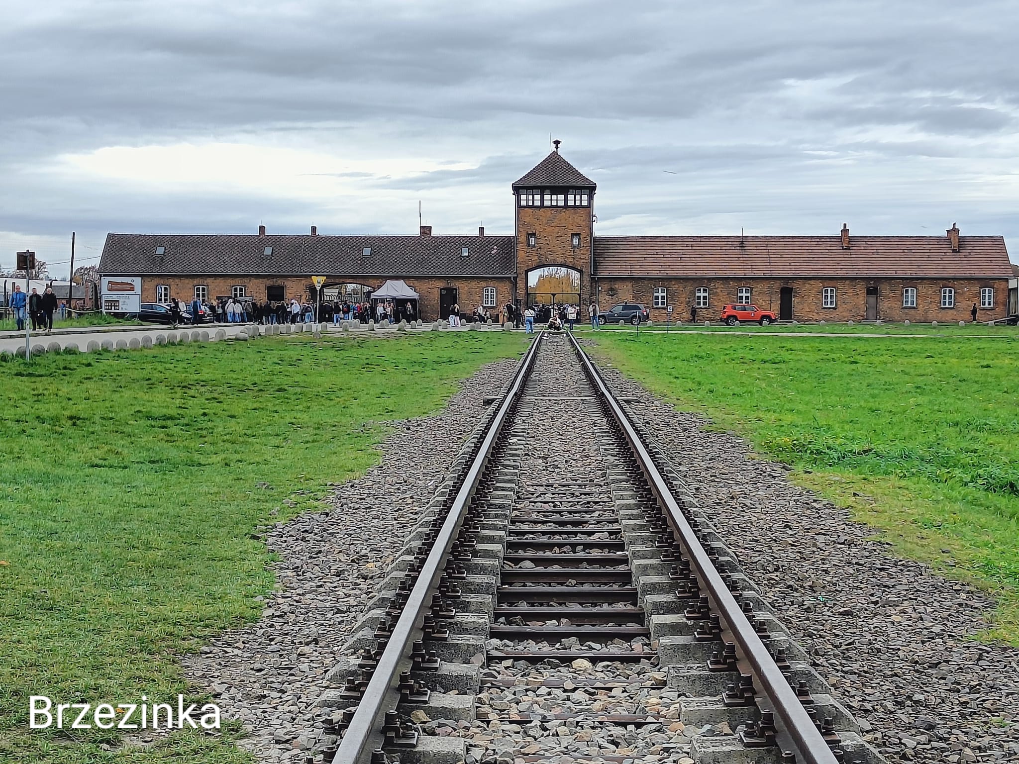 Na zdjęciu widać tory kolejowe prowadzące do byłego niemieckiego obozu koncentracyjnego Auschwitz. Tory biegną prosto w kierunku ceglanej bramy z wieżą strażniczą, przez którą widać fragment terenu obozu. Po obu stronach torów rozciągają się trawiaste pasy ziemi i niskie, murowane baraki. Niebo jest pochmurne, co nadaje zdjęciu poważny, refleksyjny nastrój.