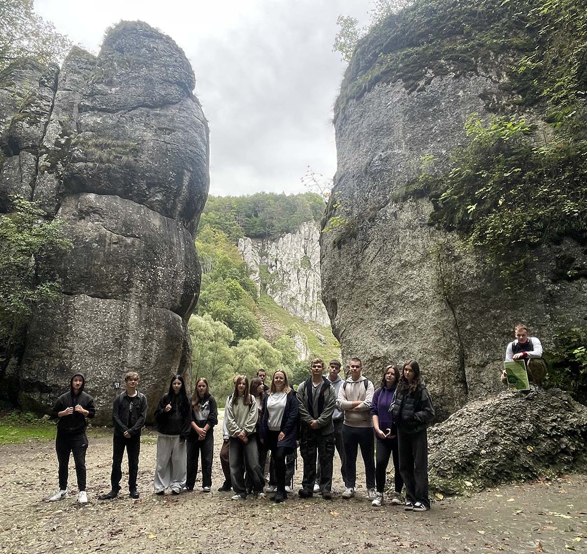 Grupa uczniów pozuje do zdjęcia na tle dwóch dużych skał. Większość osób stoi a jedna osoba kuca z boku grupy. Wszyscy ubrani są w sportowe stroje uśmiechnięci otoczeni naturalnym krajobrazem z widocznymi formacjam