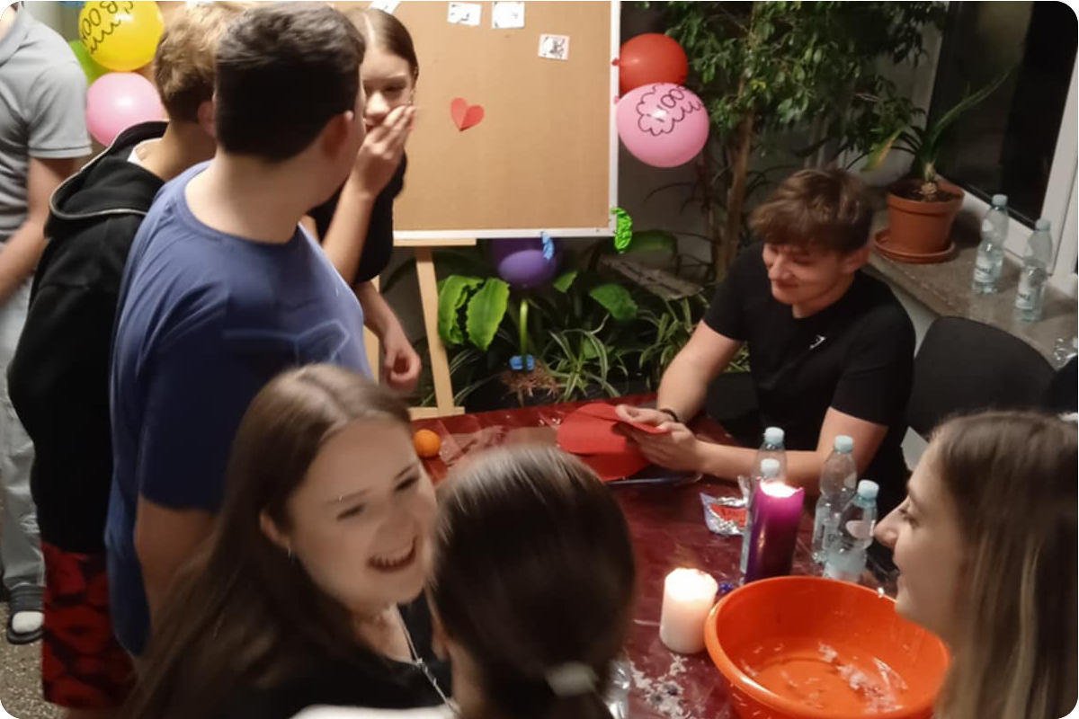 Młodzież rozmawia, śmieje się i wspólnie spędza czas bawiąc się we wróżby andrzejkowe. Na stole stoją świece, miska oraz napoje. Zdjęcie oddaje radosną, towarzyską atmosferę spotkania integracyjnego.