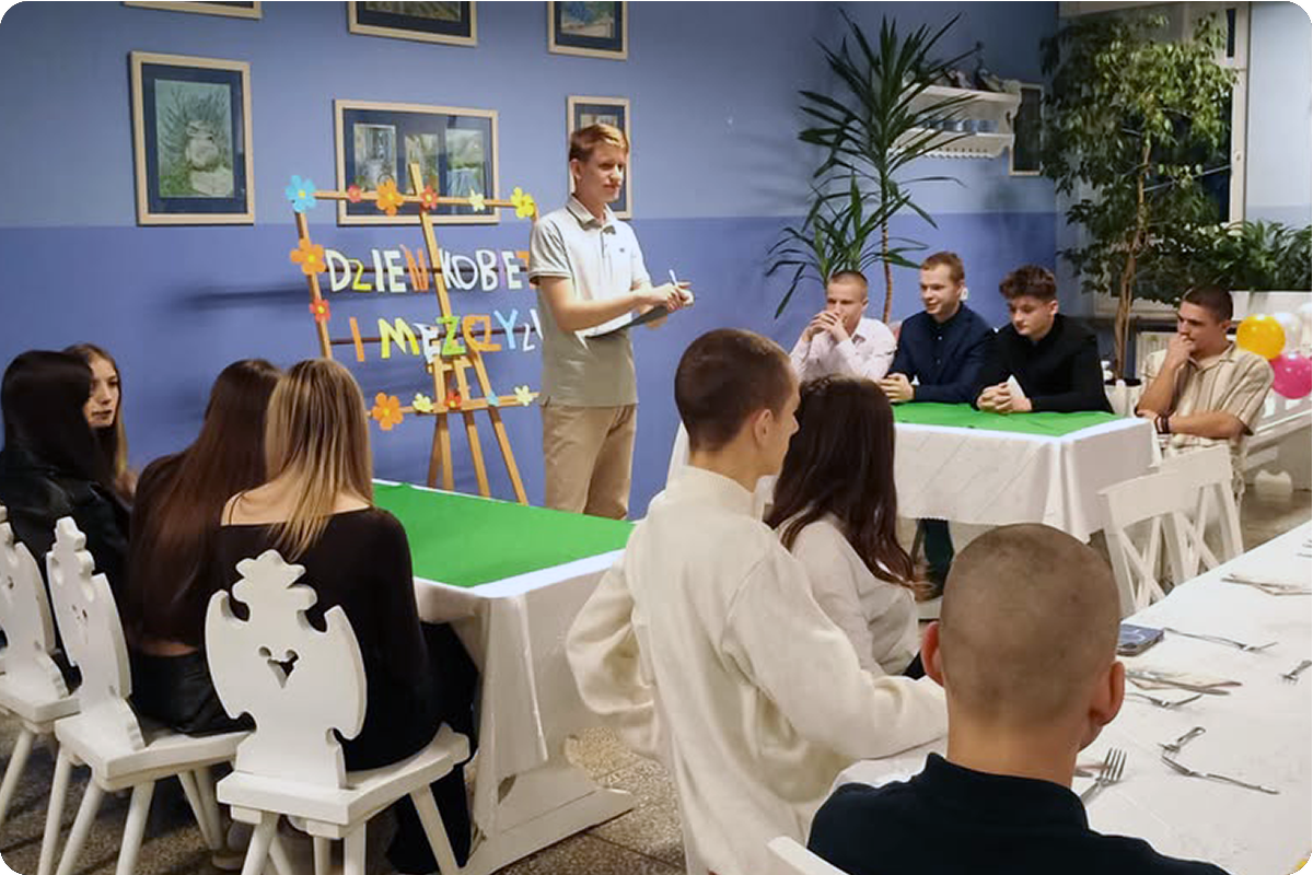 Jeden z uczestników stoi przed grupą i prowadzi quiz tematyczny, a obok niego znajduje się dekoracja z napisem „Dzień Kobiet i Mężczyzn”. Pozostali uczestnicy siedzą przy stołach i z zainteresowaniem obserwują przebieg spotkania.