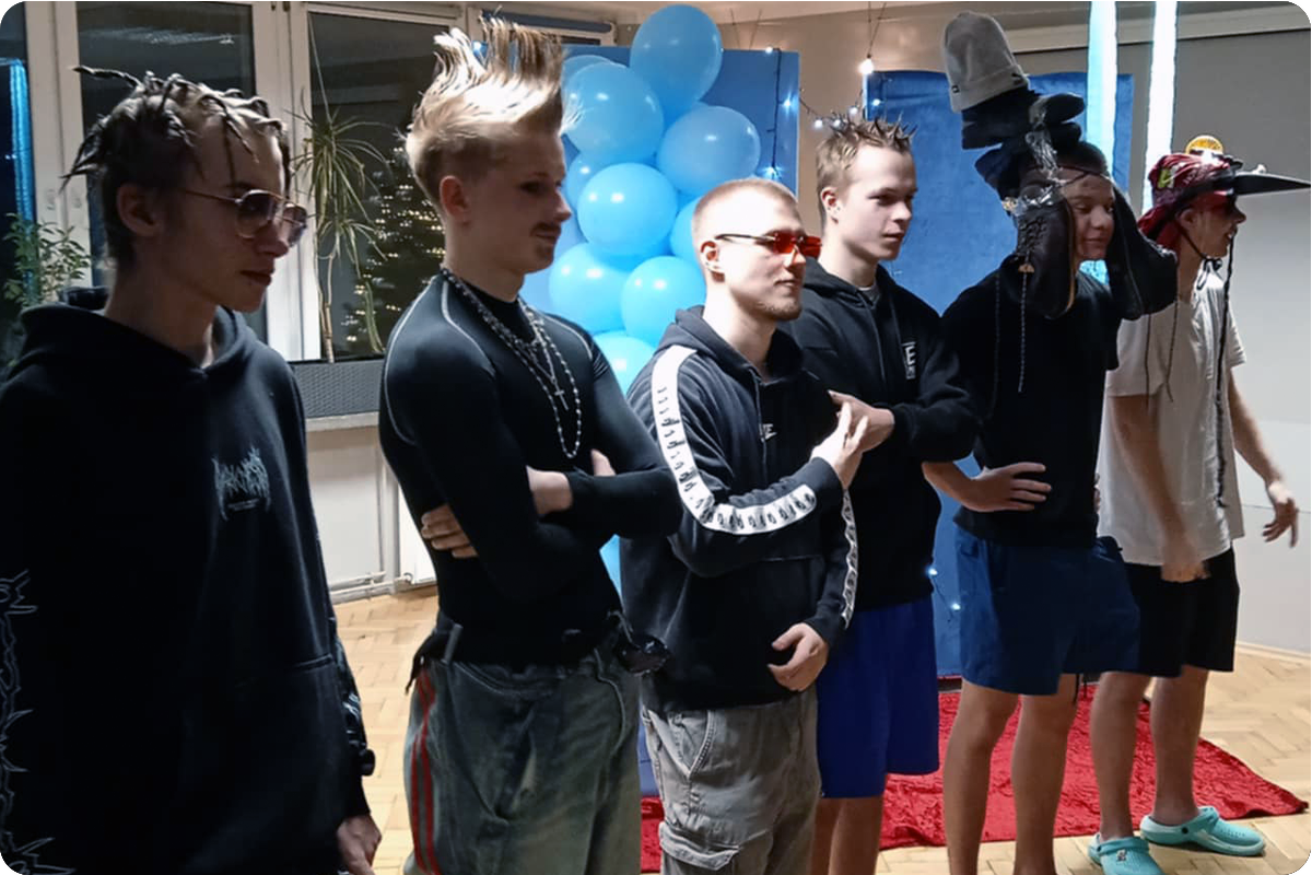 Na zdjęciu widać grupę młodych chłopaków stojących w sali udekorowanej niebieskimi balonami i ozdobami. Są ubrani w nietypowy, zabawny sposób — mają fantazyjne fryzury, okulary przeciwsłoneczne, kapcie i różne improwizowane dodatki.