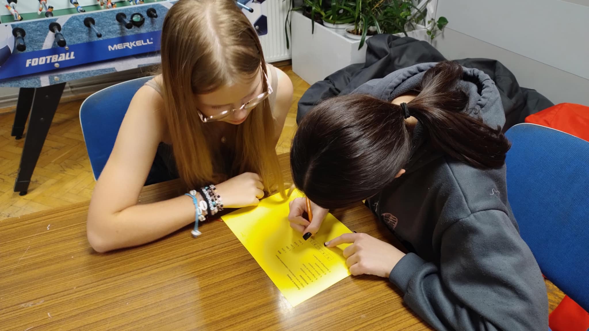 . Dwie osoby siedzą przy stole i wspólnie pochylają się nad żółtą kartką, rozwiązując zadanie lub wypełniając ćwiczenie.