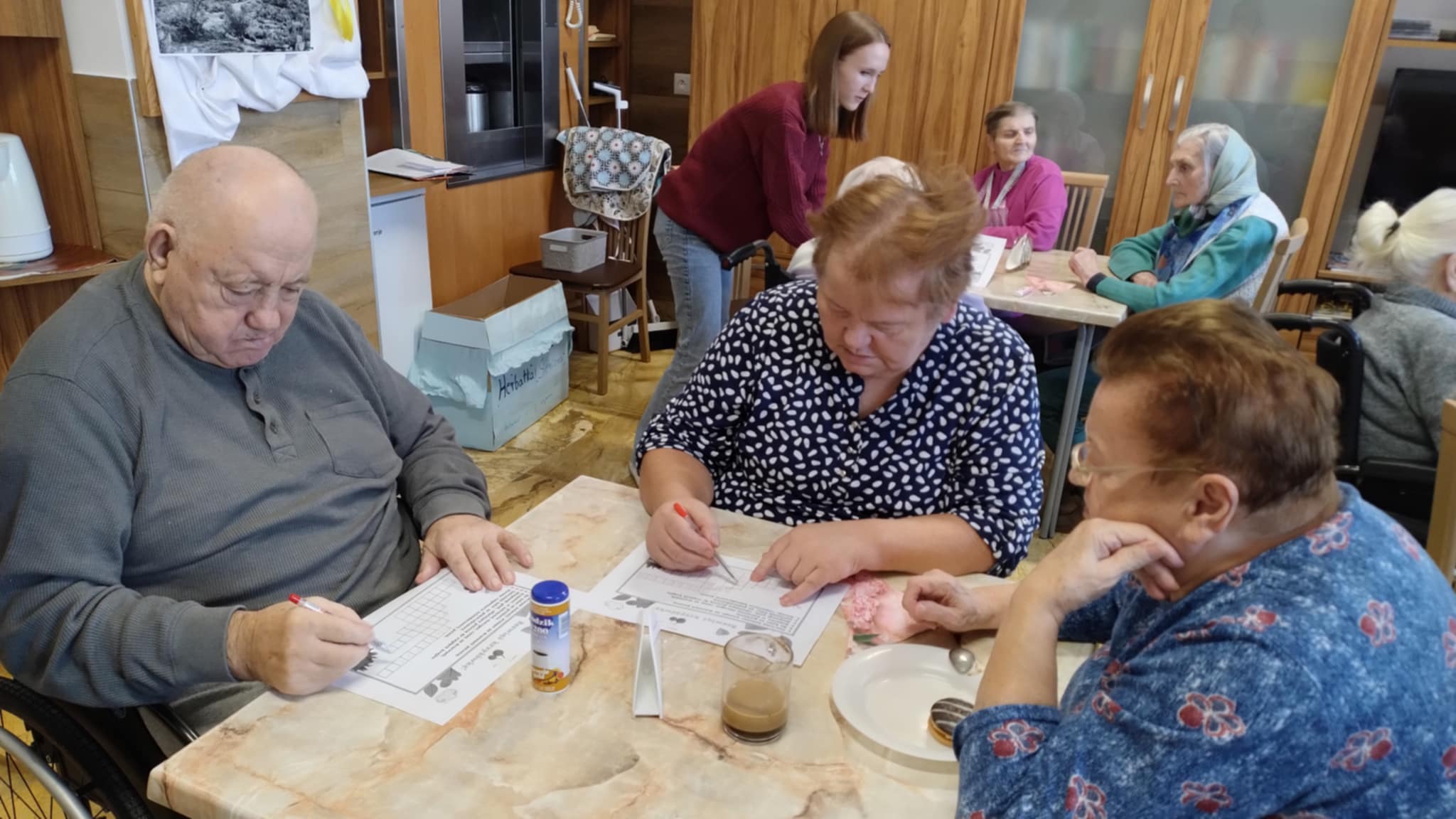 Kilka osób starszych siedzi przy stole i rozwiązuje zadania na kartkach. Mają przed sobą długopisy, ołówki i napoje. W tle widać inne osoby zajęte rozmową lub pracą przy stolikach.