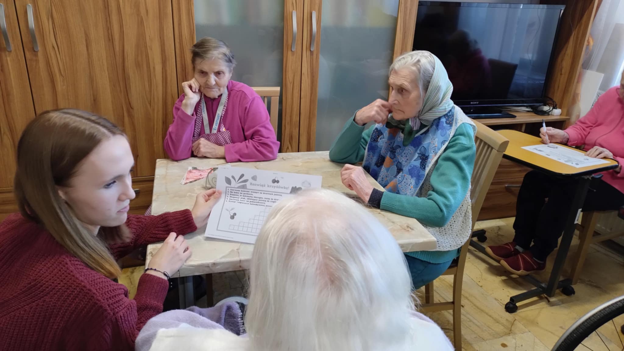 Trzy osoby starsze siedzą przy stoliku, a młoda kobieta pomaga im rozwiązywać krzyżówkę. Każda osoba ma przed sobą kartkę z zadaniem. W tle widać szafki i telewizor.