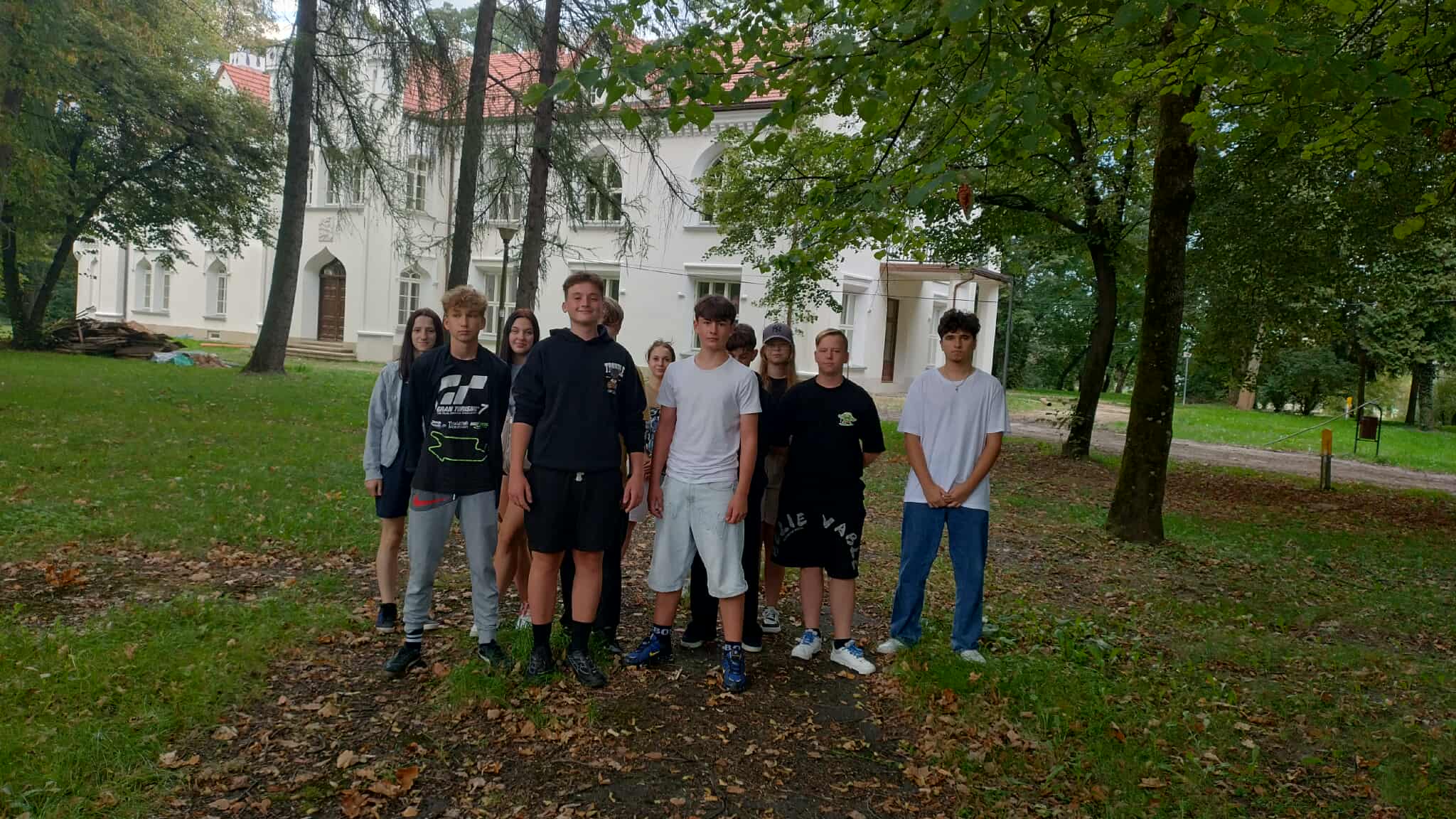 1.2	Grupa nastolatków stoi przed okazałym, białym budynkiem w otoczeniu drzew. Jest to odnowiony Pałac Dąbskich w Wojniczu. Wszyscy ustawieni są w szeregu, patrząc w stronę aparatu, w pięknej scenerii zabytkowego parku.