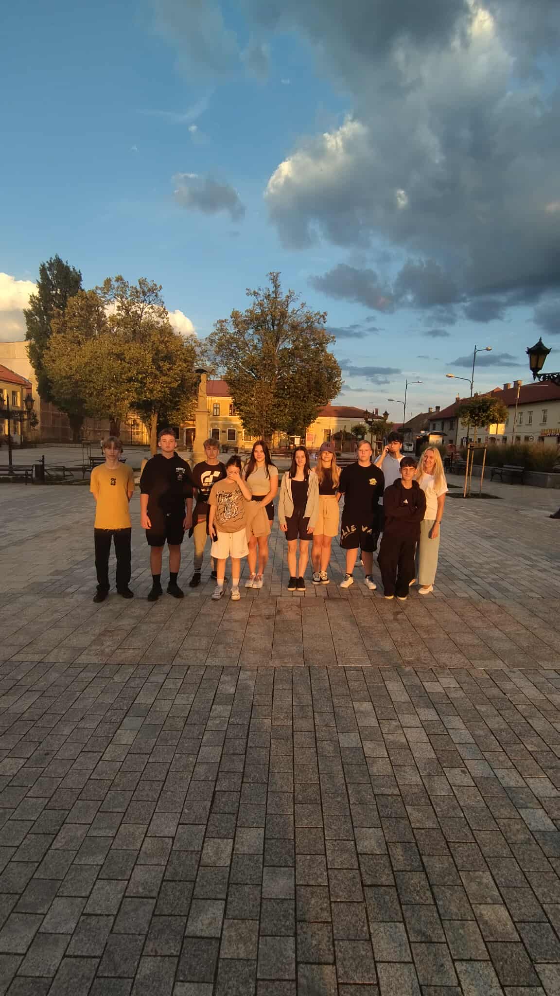 4.1	Grupa młodych osób stoi na dużym, brukowanym placu w ciepłym, złotym świetle zachodzącego słońca. W tle widać drzewa, budynki i lampy uliczne, co wskazuje na spokojne, niewielkie miasteczko. Wszyscy pozują do wspólnego zdjęcia, tworząc radosną, zgraną grupę.