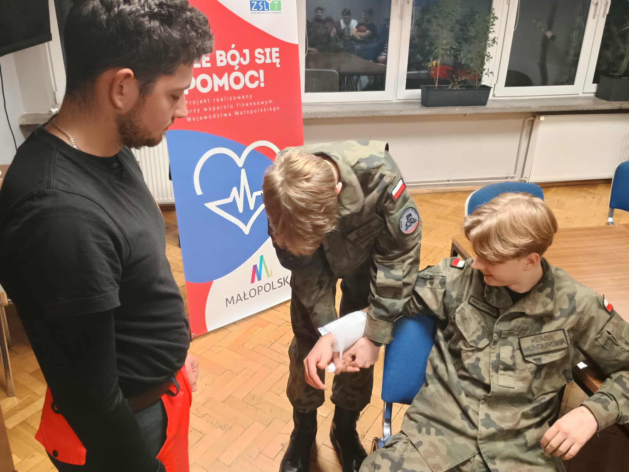 1.	Dwóch młodych ludzi w mundurach ćwiczy udzielanie pierwszej pomocy. Jeden siedzi na krześle, drugi opatruje mu przedramię bandażem. Obok stoi instruktor w czarnym stroju i czerwonych spodniach ratowniczych. W tle widoczny jest baner promujący projekt „Nie bój się pomóc!”.