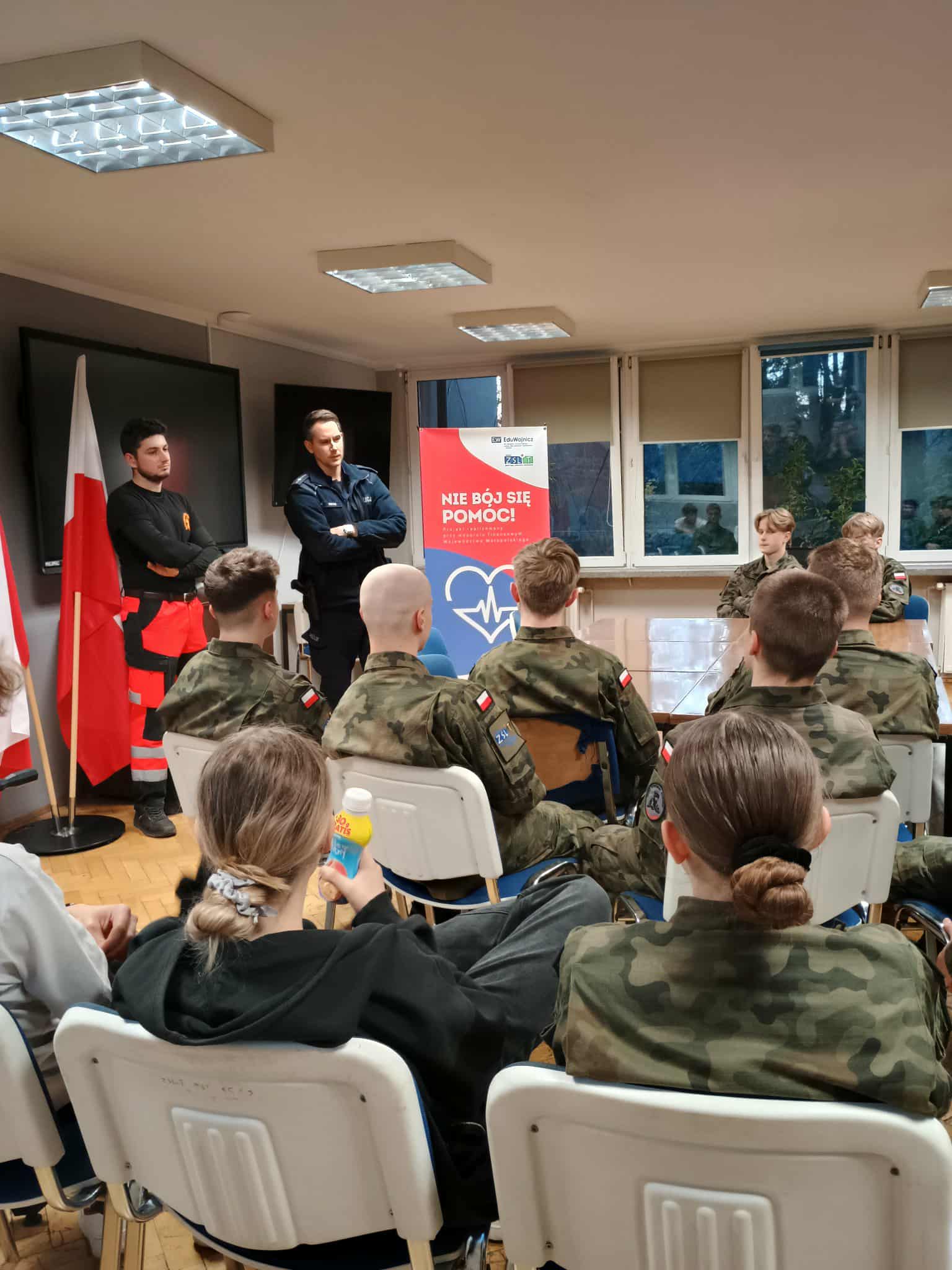 3.	Grupa młodzieży, głównie w mundurach, siedzi na krzesłach i słucha dwóch prowadzących: jednego w policyjnym mundurze, drugiego w stroju ratownika. W tle widoczny jest baner promujący projekt „Nie bój się pomóc!”.
