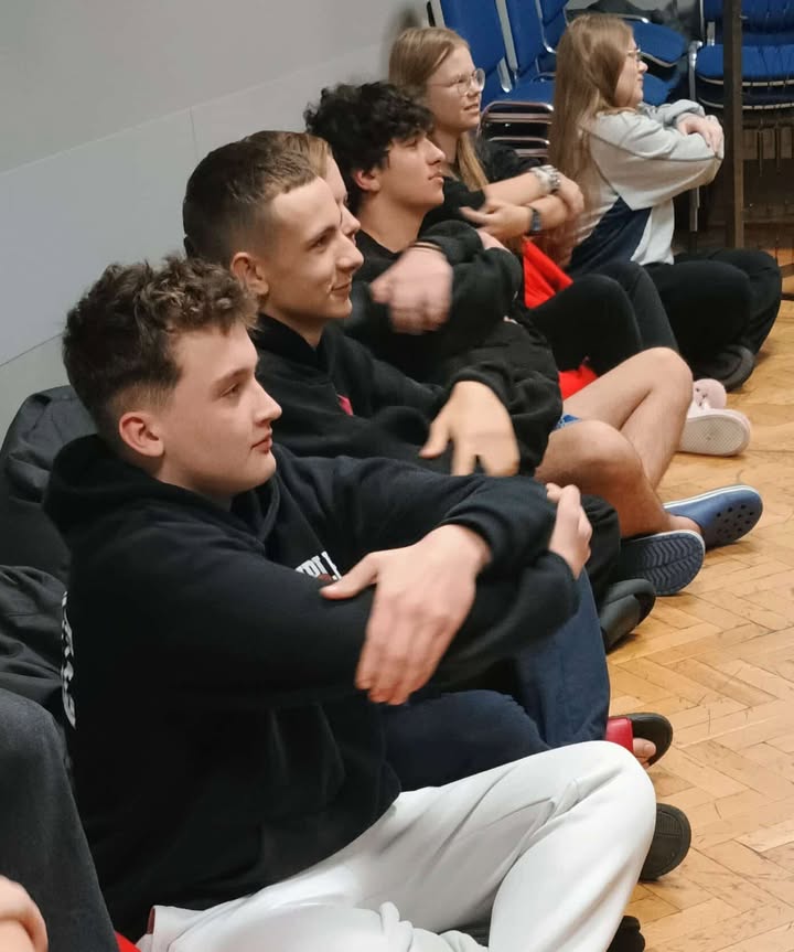 Grupa młodych osób siedzi na podłodze pod ścianą. Mają ręce skrzyżowane na klatce piersiowej i wykonują proste ćwiczenie relaksacyjne. Wszyscy wyglądają na skupionych.