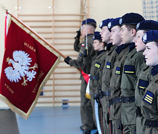 Uczniowie stoją w umundurowaniu, trzymając sztandar z wizerunkiem Jana Pawła II podczas uroczystości szkolnej.