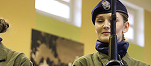 Na zdjęciu widać uczennicę podczas uroczystości, prezentującą broń w ramach pokazu lub ceremonii związanej z profilem wojskowym. Uczennica jest umundurowana i stoi z bronią, uczestnicząc w wydarzeniu.